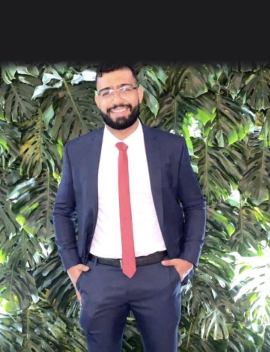 Bruno Ferraz, advogado, em pé diante de uma parede de plantas, de terno azul e gravata vermelha.