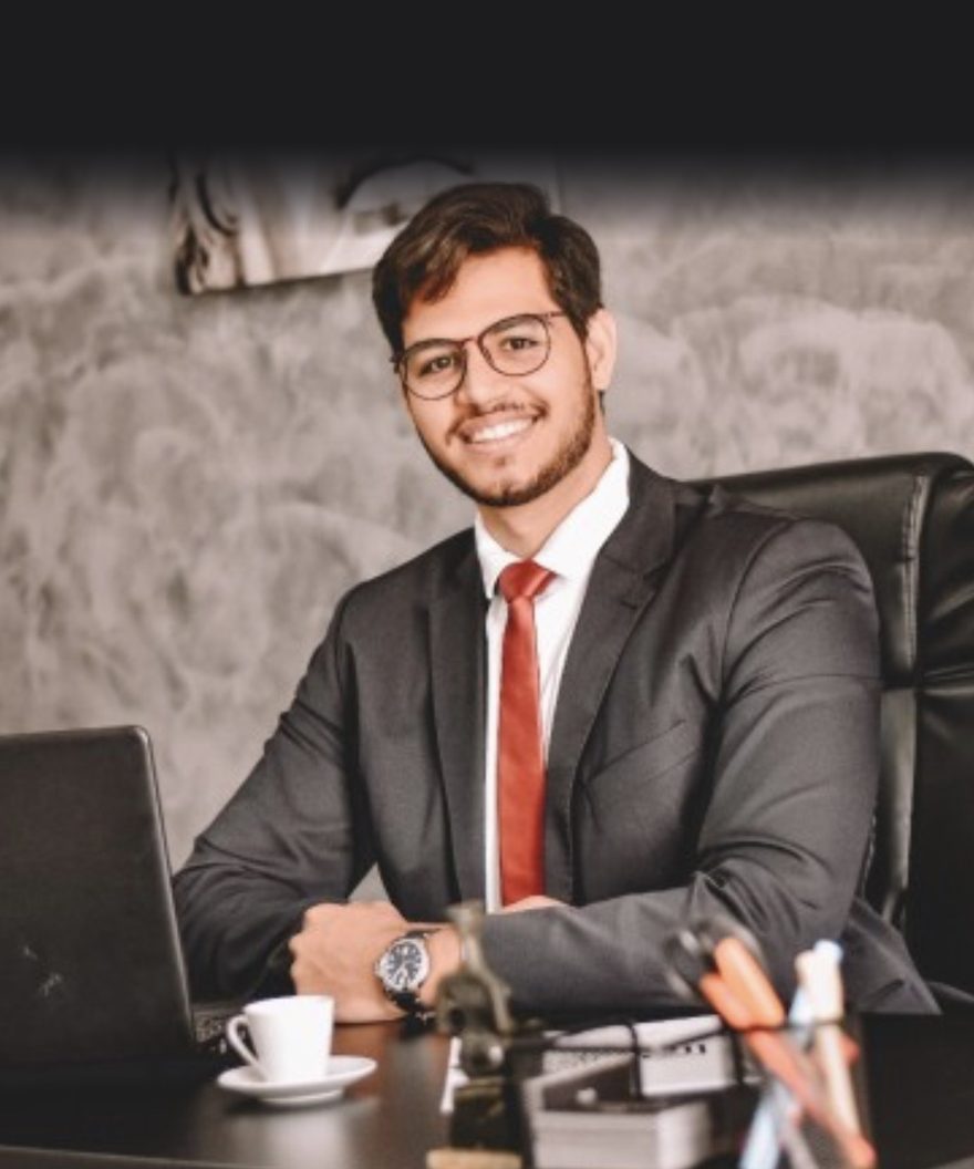 Nikson Pimenta, advogado, sentado à mesa do escritório, de terno escuro e gravata vermelha.